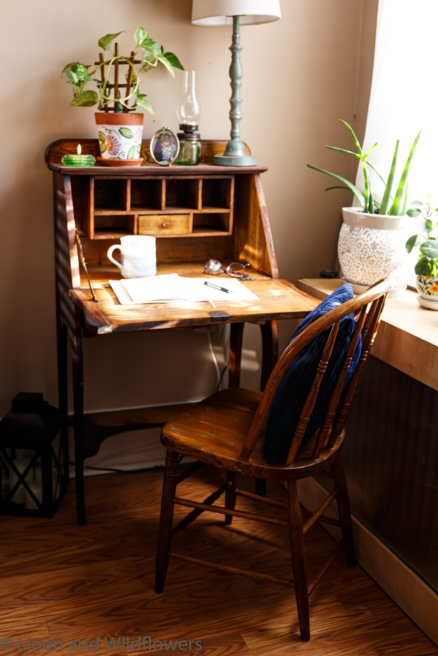 An antique secretary desk opened to write. On the open leaf, is an open note book with a pen, next to the notebook is a pair of glasses & a ble & white mug. At the top of the desk is a potho plant in a hand painted terracotta planter, an candle votive holder with a hurricane lamp shape, a photo of a grandmother & granddaughter wmbracing in a hug. Next to the planter is a Fenton candle holder with a lit candle. Next it the desk is a covered radiator, on top of the radiator cover is houseplants basking in the sunny window.