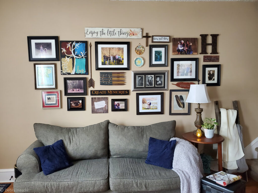 A large wall gallery of family photos, art, and memorabilia over a couch.  On the couch is two blue suede pillows & a fuzzy grey throw blanket draped over the arm of the couch. On the end table is a wood base lamp with a white lamp shade, a Fenton candle holder & a milk glass panter with a potho plant in it.