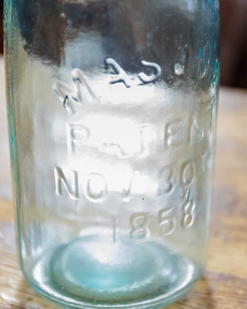 Close-up of a vintage glass jar with embossed text reading "MASON PATENT NO 30 1858." The jar is light blue, suggesting an antique, rustic feel.