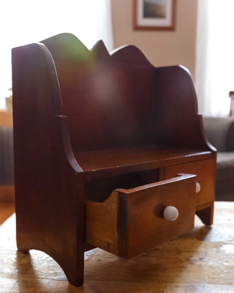 A small, vintage wooden cabinet with a scalloped back, featuring two drawers with white knobs, one slightly open, sits on a wooden table in a cozy room.