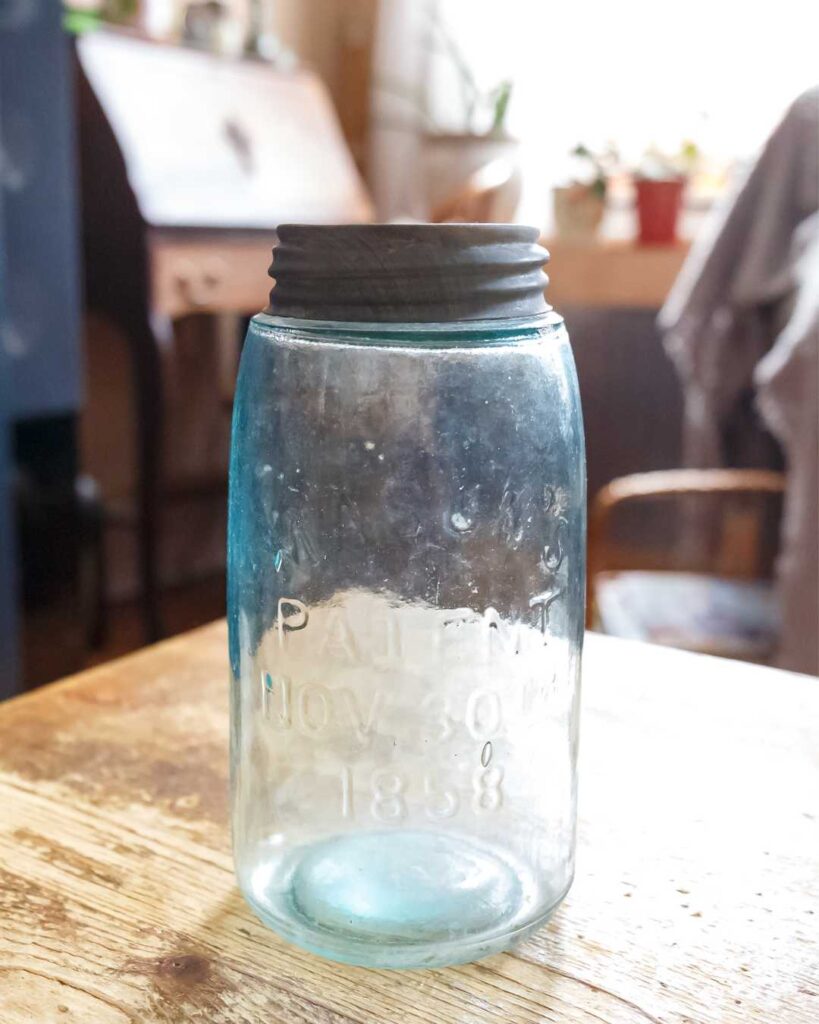 Antique blue glass jar with metal lid on a wooden table. "Patent Nov 30th 1858" embossed on the front. Warm, nostalgic ambiance with a soft background.