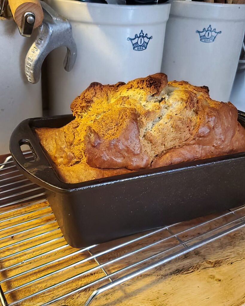 Golden loaf of homemade bread cooling in a cast iron loaf pan on a wire rack.