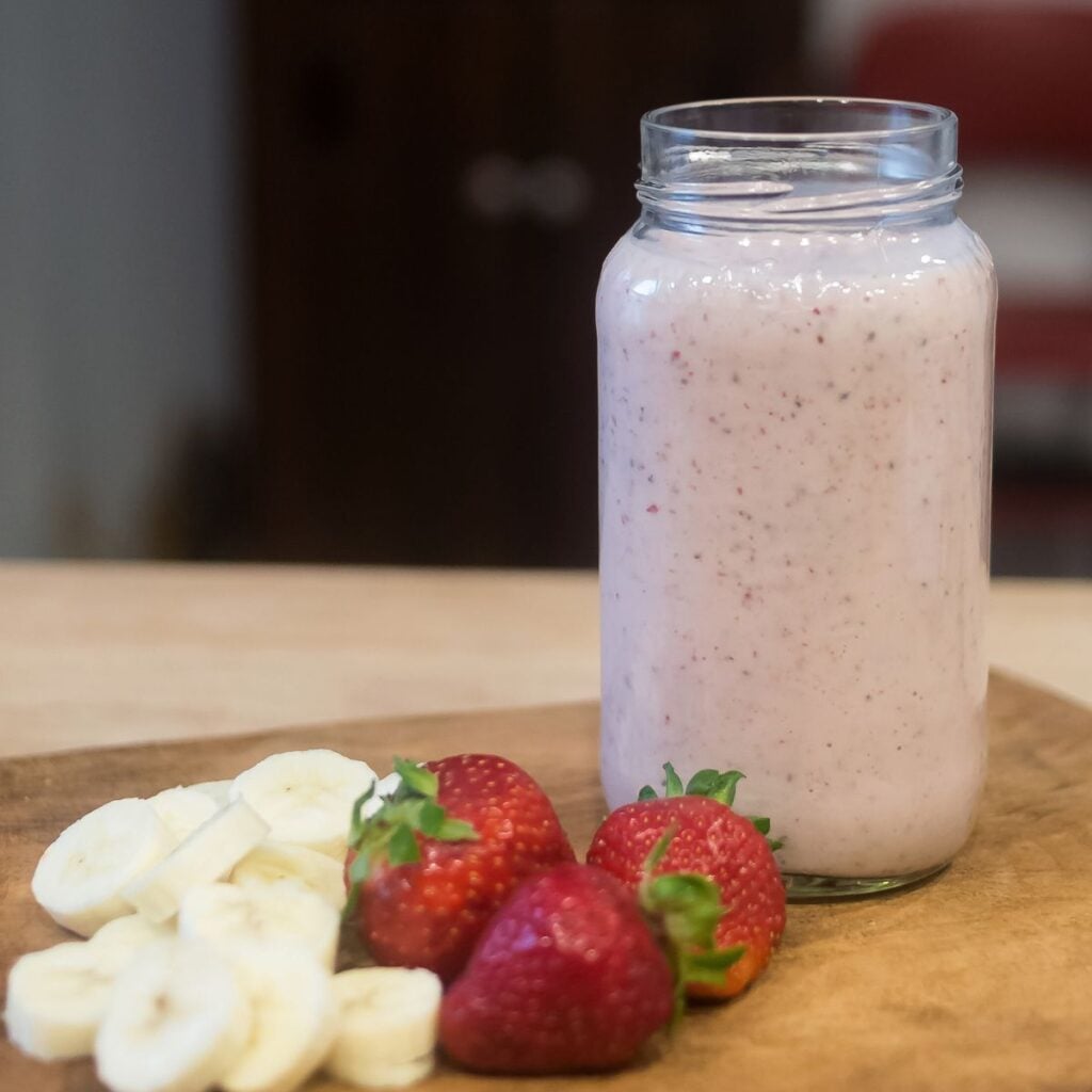A feature image for Strawberry Banana Smoothie Recipe-www.LinenandWildflowers.cm Photo Description: A delicious strawberry banana smoothie in a mason jar next to strawberries and cut up sliced bananas on a cutting board.