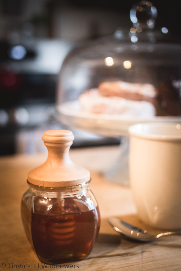 Honey Jar with a wood top next to a white mug, spoon & a clear glass cake stand.