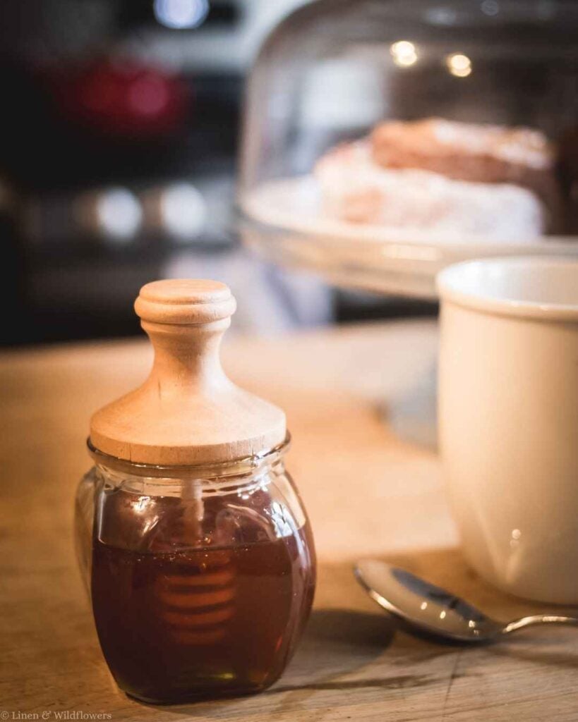Close-up of a wooden honey dipper resting in a glass jar next to a white tea mug in a farmhouse kitchen.