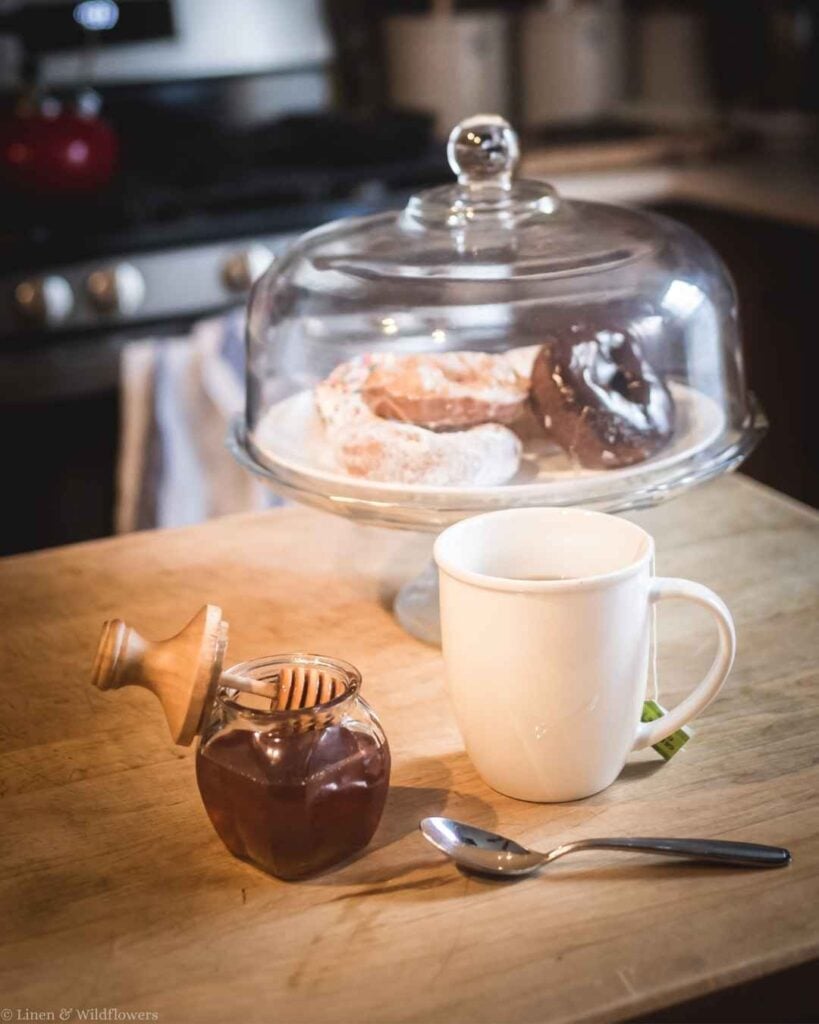 Glass honey jar with wooden dipper lid on a wood counter beside a tea mug and donuts under a glass cloche.