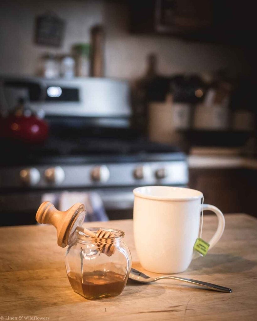 Rustic glass honey jar with a turned wood lid sitting on a wooden countertop with soft kitchen bokeh.