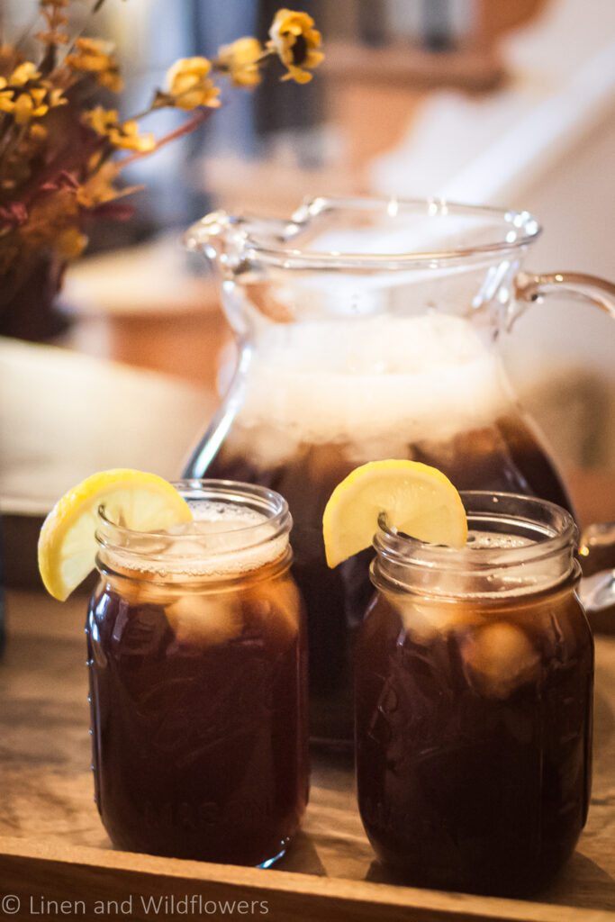 A pitcher of homemade ice tea on a wooden tray with two mason jars with iced tea ^ lemon slices.