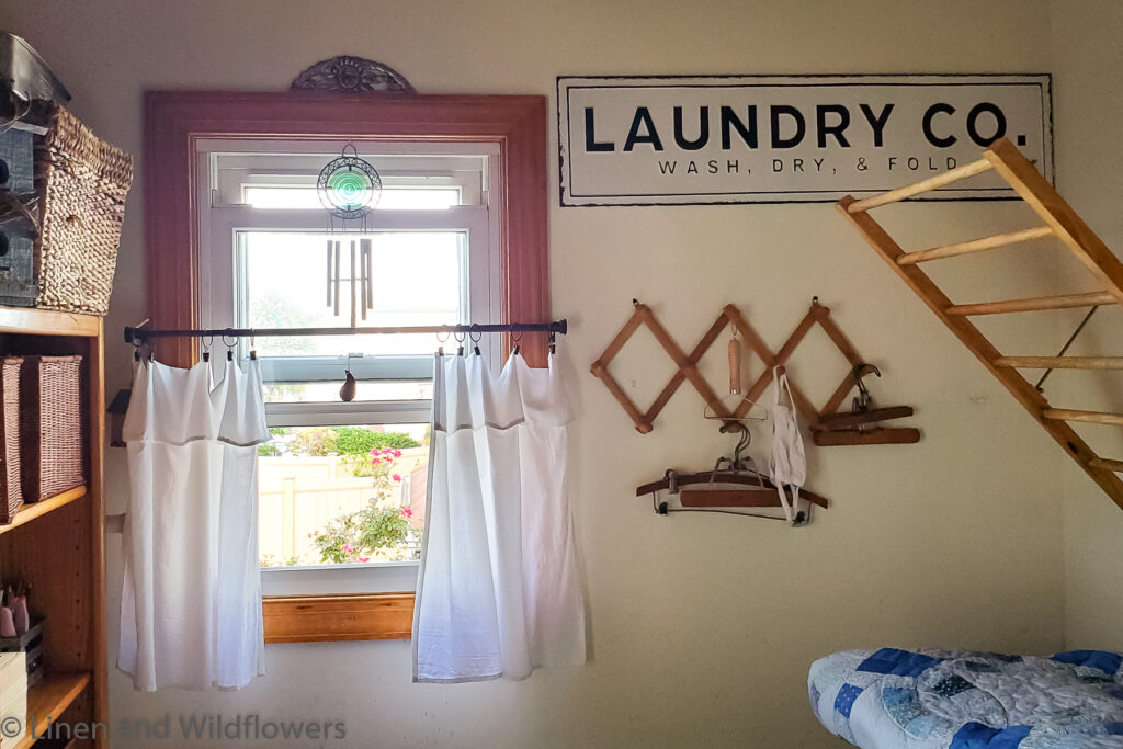 No Sew Tea Towel Curtains & wind chimes hanging on a laundry room window. On the left is a bookcase with baskets used for storage. On the right is a Laundry sign, a peg rack with antique hangers & mask & a drying rack.