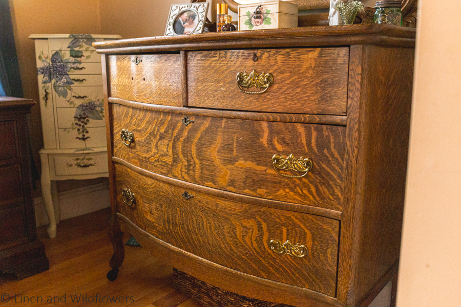 Tiger wood Victorian Dresser with its original gold hardware & key locks.  Under the dresser is a wicker basket for storage. In the corner next to it is a floral painted large jewelry box.