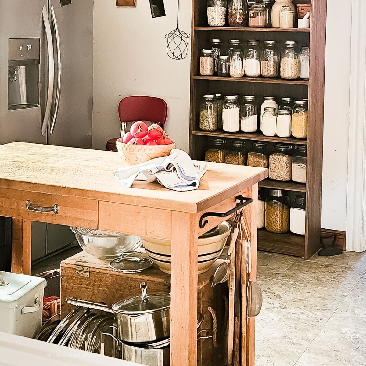 Warm kitchen scene with a wooden island, bowls, and pots. A basket of tomatoes sits on a towel. Shelves with jars of grains and spices create a cozy, organized vibe.