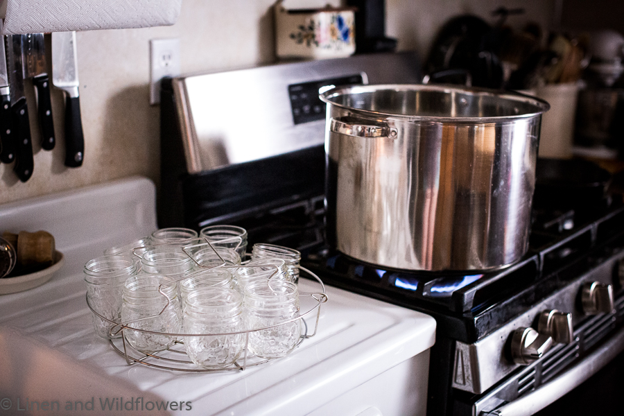 Simple Water bath Canning-A canning pot on a lit stove & a rack of mason jars on sink drainboard next to stove.