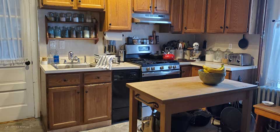 A dated kitchen with a two tiered shef above the kitchen sink filled with mason jars. A butcher block with a bou=wl of apples & bananas. A small dishwasher & countertops with crocks of utensils, toaster, kitchen aid mixer, cutting board & canisters.