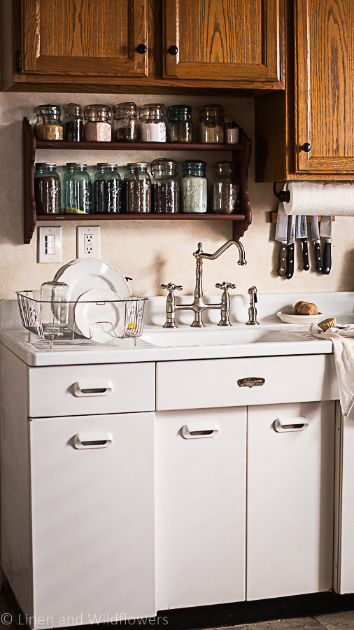 How to Organize Under a kitchen sink a vintage 1950s white sink with metal cabinets and a fance vintage faucet. On the drain board to the left is a metal drainer on the drainboard with ironstone dishes drying, over to the left is a magnet on the wall with knives & a papertowel holder. Above the sink is a burgundy two tiered shelf with a variety of mason jars filled with dry goods. 