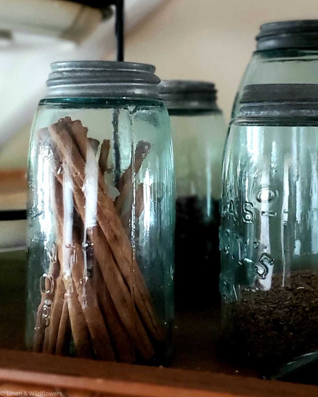 Close-up of vintage glass jars on a wooden shelf, filled with cinnamon sticks and spices. The rustic setting conveys a warm, nostalgic feel.
