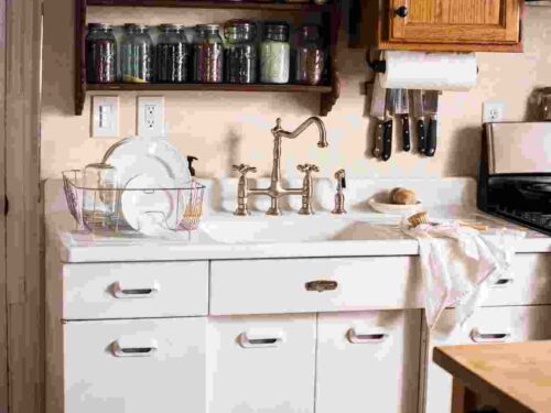 a vintage 1950s white sink with metal cabinets and a fance vintage faucet. On the drain board to the left is a metal drainer on the drainboard with ironstone dishes drying, over to the left is a magnet on the wall with knives & a papertowel holder. Above the sink is a burgundy two tiered shelf with a variety of mason jars filld with dry goods.