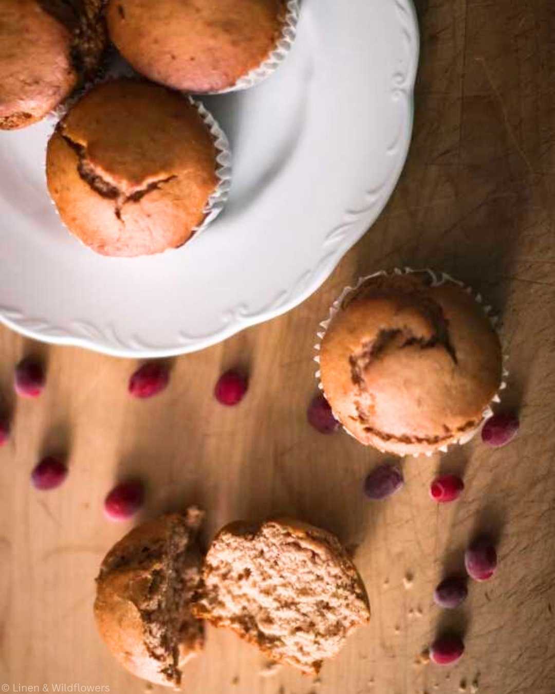 Freshly baked homemade cranberry sauce muffins on a white ironstone dish with scattered cranberries on a wooden surface. A broken muffin displays a soft, moist interior. Cozy and inviting.