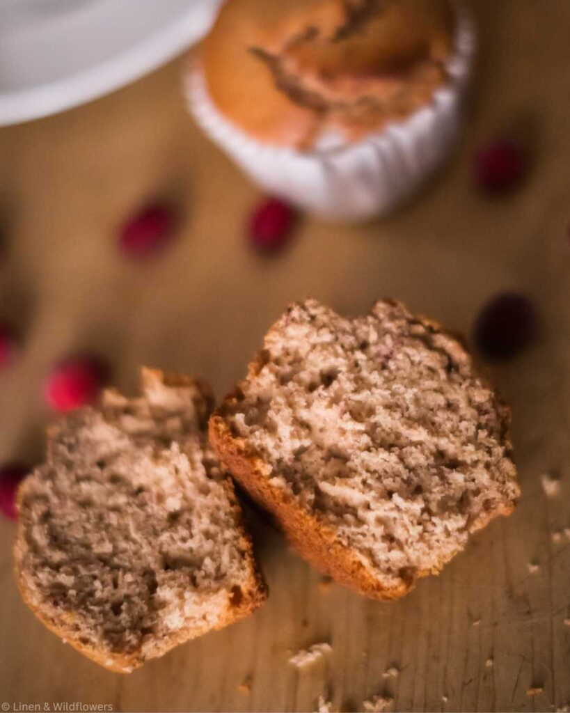 Cracked cranberry muffin top with a broken half atop a wooden surface, surrounded by scattered red berries, conveying a cozy, homemade feel.