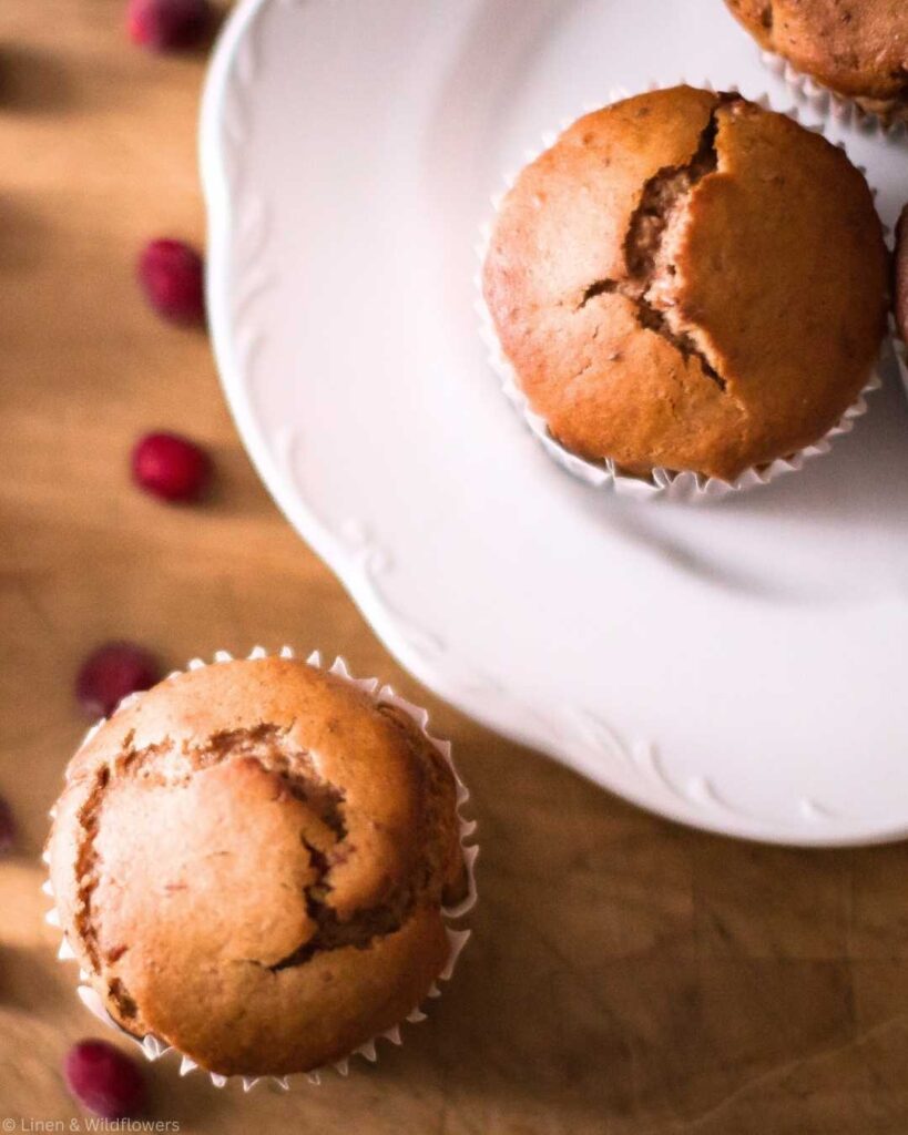 Two golden-brown cranberry sauce muffins in liners are displayed on a white ironstone dish and wooden surface with scattered cranberries, conveying a cozy homemade feel.