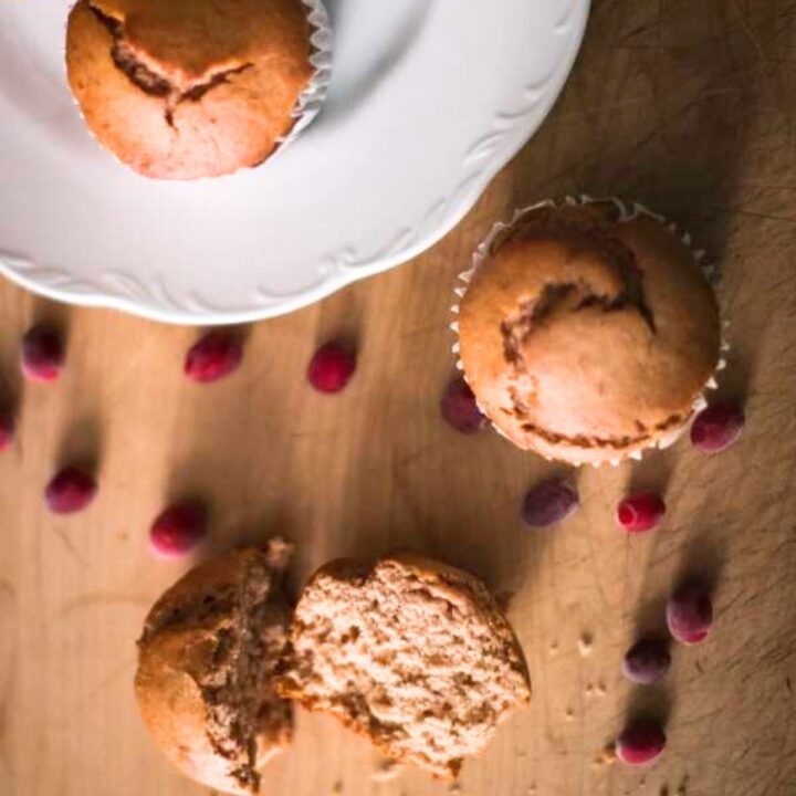 A split cranberry sauce muffin on a wooden table with scattered cranberries radiates a cozy and inviting atmosphere. Another muffin in a wrapper is partially visible.