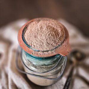 A jar filled with homemade hot chocolate mix, nestled in a vintage mason jar, rests on a fabric surface. A small scoop is beside it, suggesting a culinary or baking setting.