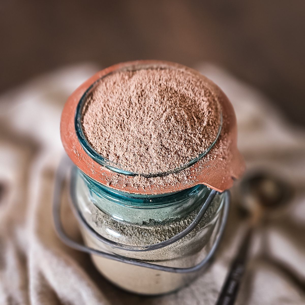 A jar filled with homemade hot chocolate mix, nestled in a vintage mason jar, rests on a fabric surface. A small scoop is beside it, suggesting a culinary or baking setting.