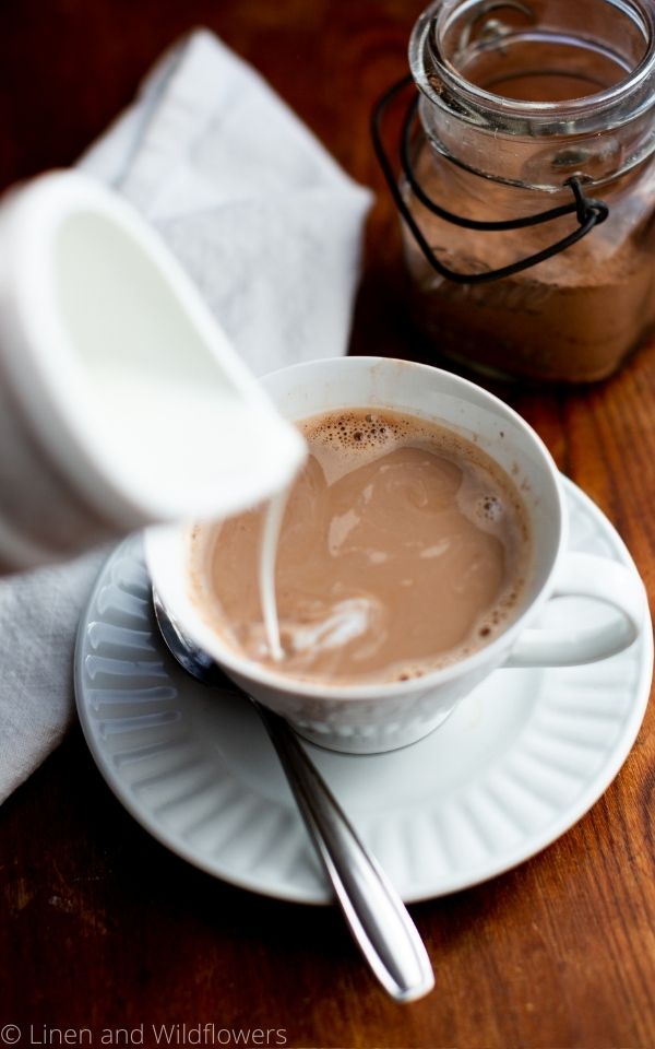 creamer being poured into a cup of hot mocha resting on a saucer with a spoon.