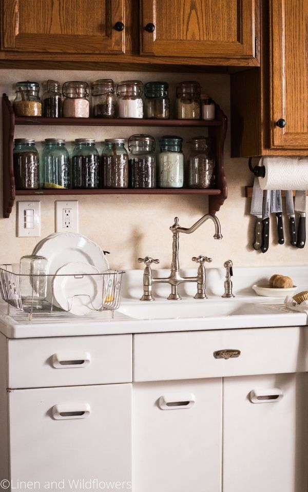 16 Useful Kitchen Tips-vintage sink with a shelf above with vintage mason jars of dy goods. A scrub brush on sink drain board & a dish & in dish rack. Above the sink to the right of five kitchen knives on the wall held by a magnet.