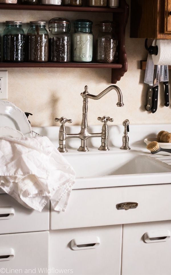 16 Useful Kitchen Tips-vintage sink with a shelf above with vintage mason jars of dy goods. A scrub brush on sink drain board & a dish & tea towel on dish rack. Above the sink to the right si three kitchen knives on the wall held by a magnet.