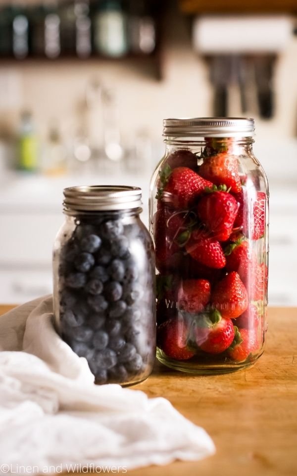 a quart mason jar of blueberries & a 2 quart jar of strawberries on a butcher block with a yea towel/