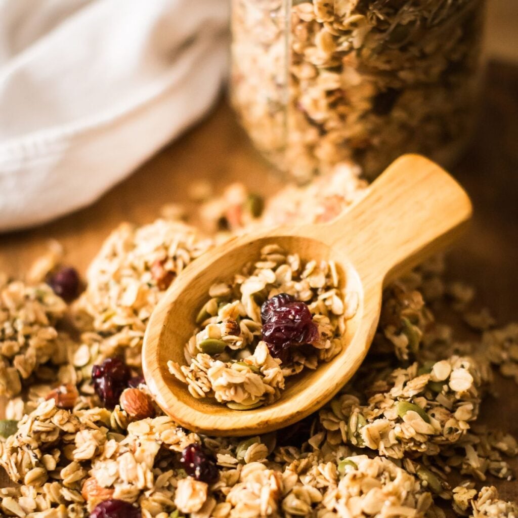 Wooden spoon scooping granola mix with oats, almonds, and cranberries on a wooden table. The warm, rustic setting conveys a cozy, homemade feel.