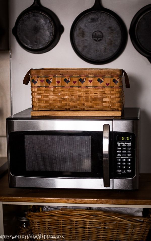 a stainless steel microwave on a cabinet with a basket on top & cast skillets hanging above the basket & microwave,