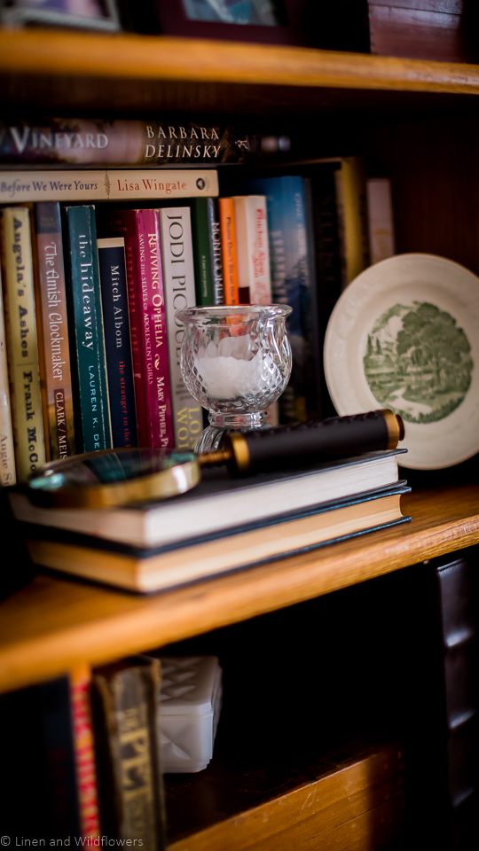 Using Inexpensive Ideas to Update Your Space is a bookcase filled with a books, a vintage mini green & white plate on a stand, antique magnifying glass & a candle votive holder.