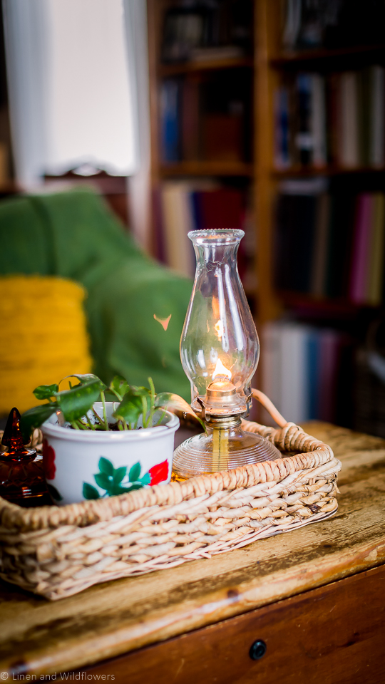 a wicker basket tray with a oil lamp, plant in a floral planter & a orange candy dish. In the background is a bookcase filled with books & decor items.