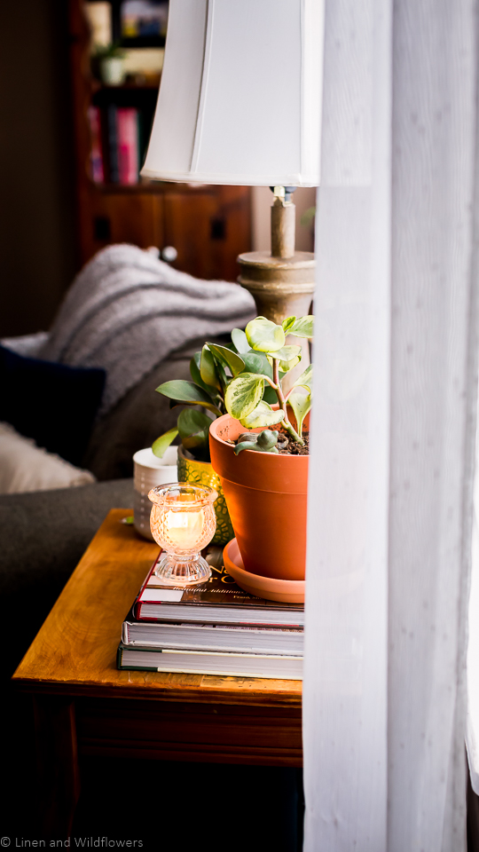 An end table next to a chair with pillows & a throw with a stack of decor books and a lit candle in a clear votive holder next to plants.