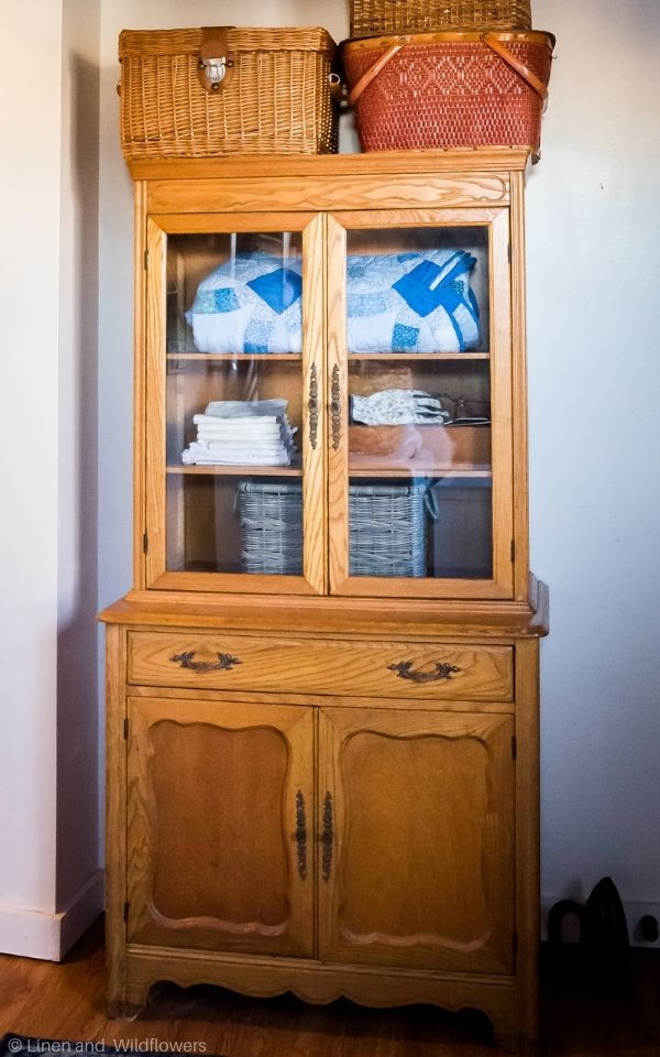 a vintage almond maple stepback cupboard used for holding linens & supplies for a laundryroom and large baskets sitting on top.