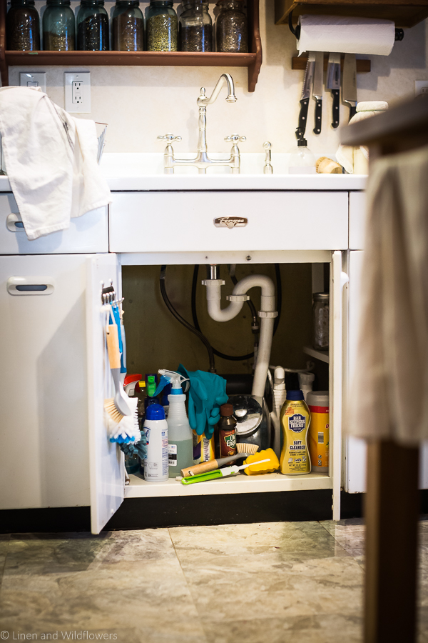 the cabinet doors are opened under the vintage sink with cleaning products that are in a disarray. How to Organize Under a kitchen sink willbe needed to get htis cabinet in order,