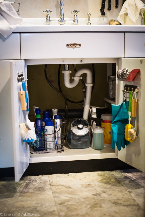 How to Organize Under a kitchen sink with hooks & caddy on the doors to hold cleaning brushes & gloves, a basket inside cabinet holding cleaning supplies, a glass spray bottle with homemade cleaning solution and a container holding brillo pads. Next to it is a cleaning spray, disinfected wipes & another all-purpose cleaner.