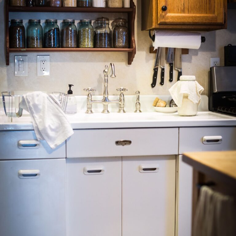Cozy kitchen with white cabinets, vintage sink, and faucet. Shelves hold jars of grains and spices, creating a warm, rustic atmosphere.