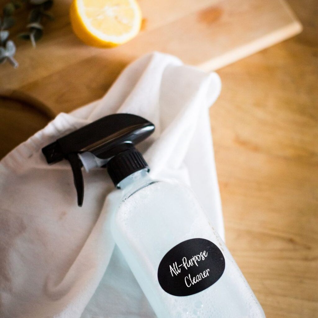Homemade all-purpose cleaner in a glass bottle lays on a towel next to a cutting board & sliced lemon.