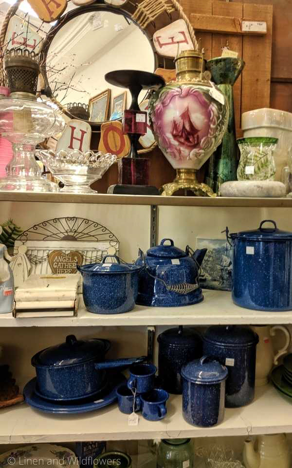 A thrift store with shelves of items for sale, including blue enamelware, oil lamp, dishes & so much more,