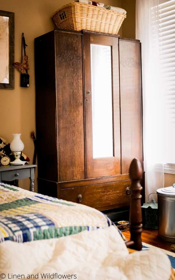 An antique armoire in a bedroom with a large basket of quilts that sits on tp next to a blue bed table with a vintage alarm clock & lantern lamp. 