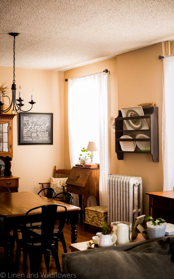 The Art of Styling Your Home-A dining room includes a variety of furniture including a diningroom table, secretary desk, chair, side table with decor. The room also has its aoriginal radiator from 1885, above it is a plate plate rack with serving platters.
