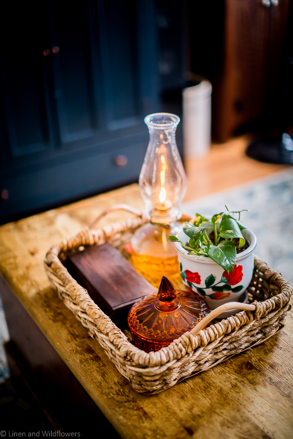 A wicker tray on a blanket chest curated with a wooden box, an antique oil lamp. amber colored candy dish & a floral planter with a potho plant. 