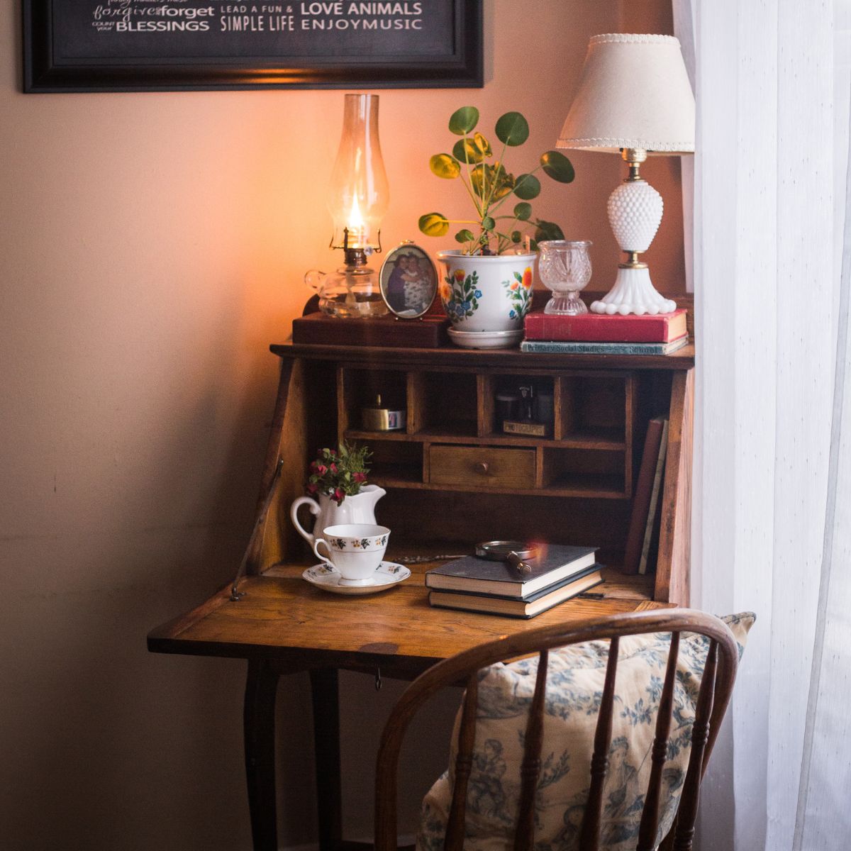 A antique secretary desk in the corner of the room decorated with an oil lamp, small gold photo frame, a houseplant, and two old books with a candle holder & a milkglass lamp. Inside the desk has 2 books with a magnifying glass on top, next th that is a vintage tea cup & saucer & a ironstone creamer with a dried bouquet of mini roses. Above the desk is a black & white print with last name surrounded with family rules.