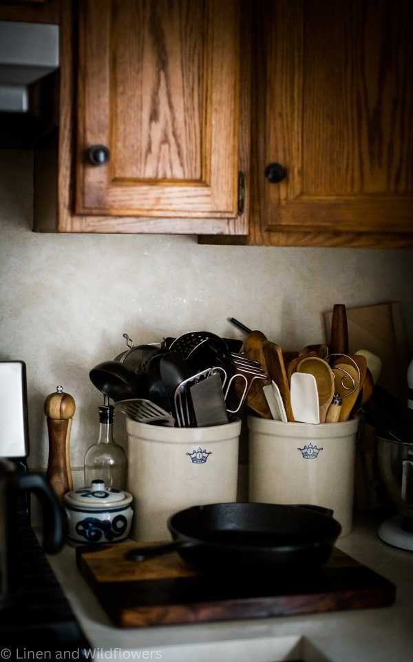 Two crocks on the counter filled with cooking utensils. On the counter in front of the crocks is a wood cutting board & a cast iron skillet, blue & white salt cellar, pepper grinder & oil bottle.