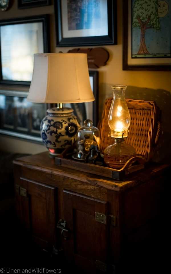 How to Decorate with a oil lamp is easy to do. A vignette curated on a wood tray of a oil lamp, cloche filled with shells & a cast iron liberty bell sitting by a blue & white lamp with a white shade on an antique ice box.