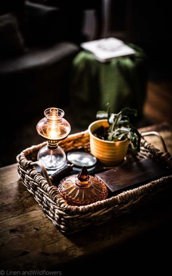 A wicker tray with a small lit  oil lamp, amber candy dish, Christmas cactus plant in a mustard yellow planter & a wooden rectangular box. Next to the little oil lamp is antique magnifying glass.