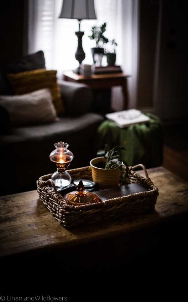 A wicker tray with a small lit  oil lamp, amber candy dish, Christmas cactus plant in a mustard yellow planter & a wooden rectangular box. Next to the little oil lamp is antique magnifying glass. In front of the wicker tray on a coffee table is a chair with cozy pillow, next to it is a end table with home décor books, two plants, candle holder, lamp & a coffee cup. in front of the table is a small ottoman with a green throw blank, open book & glasses.
