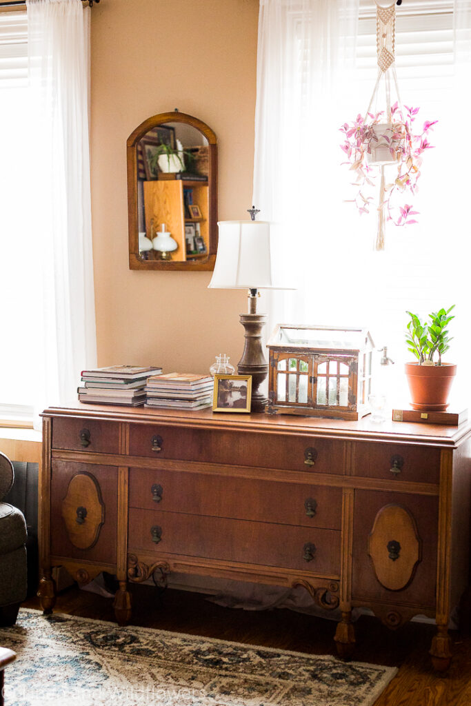 5 Uses for an Antique Sideboard- A beautiful antique side in front of a window in the livingroom styled with stacks of books, a gold landscpae frame, oil lamp, terraium with houseplants, candle, vintage spritzer & another house plant resting on an old cigar box.